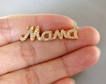 Puede incluir: Un colgante dorado con la palabra "Mama" escrita en cursiva. Las letras están incrustadas con pequeños cristales transparentes. El colgante se muestra sobre un fondo neutro.