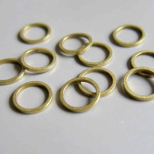 May include: A collection of small, round, brass rings scattered on a light-colored surface. The rings have a simple, minimalist design and a warm, golden hue. These rings could be used for jewelry making or other craft projects.
