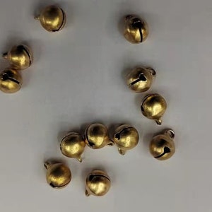 Könnte beinhalten: Eine Ansammlung kleiner, runder, goldener Glocken auf einer weißen Oberfläche. Jede Glocke hat eine kleine Schlaufe oben zum Aufhängen. Die Glocken haben eine glänzende, metallische Oberfläche und einen Schlitz an der Seite.