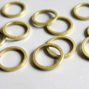 Puede incluir: Una colección de pequeños anillos redondos de metal color latón. Los anillos están dispersos sobre una superficie blanca, con algunos superpuestos. Los anillos tienen un aspecto ligeramente texturizado y un tono cálido y dorado.