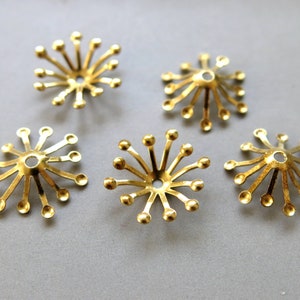 May include: Five gold-colored metal flower-shaped embellishments with a circular hole in the center. Each embellishment has 12 points that extend outward from the center.