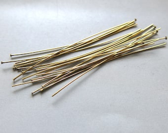 May include: A collection of gold-colored head pins, each with a straight, slender shaft and a small, flat head. The pins are scattered on a light gray surface, ideal for jewelry making and crafting projects.