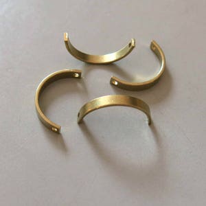 May include: Four curved, brass-coloured metal pieces with small holes at each end. The pieces are arranged on a light grey surface, casting soft shadows. The objects appear to be components or hardware.