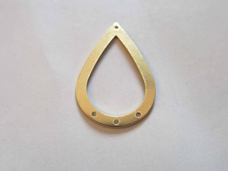 May include: A teardrop-shaped brass jewelry finding with a smooth, matte finish. The pendant has a cutout center and three small holes at the bottom, and one at the top. The metal has a warm, golden hue.