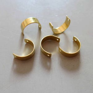 May include: Five gold-colored metal hoops with a rounded shape and a small hole on each side. The hoops are arranged in a semi-circle on a light gray surface.