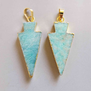 May include: Two arrow-shaped pendants with a gold-colored trim and bail. The pendants have a light blue and white marbled appearance. The pendants are displayed on a light gray background.
