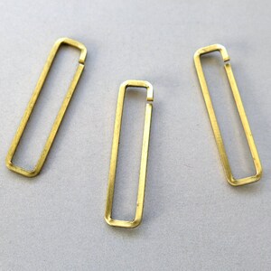 Könnte beinhalten: Drei goldfarbene Metallringe, jeder in Form eines Rechtecks mit abgerundeten Ecken. Die Ringe sind an einer Seite offen und haben eine leicht gekrümmte Form.