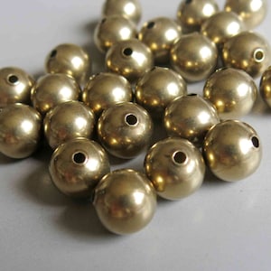 May include: A collection of small, round, metallic gold beads. Each bead has a small hole drilled through the center. The beads are scattered, with varying light reflections on their surfaces. Ideal for jewelry making or craft projects.