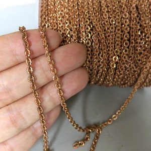 Puede incluir: Un carrete de cadena de color dorado, con un tramo de cadena drapeado sobre una mano. La cadena es de estilo cable delicado.