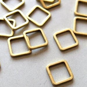Könnte beinhalten: Eine Ansammlung kleiner, quadratischer, goldfarbener Metallringe auf einer hellgrauen Oberfläche. Die Ringe haben abgerundete Ecken und eine hohle Mitte, was darauf hindeutet, dass sie dekorative oder funktionale Elemente für Bastelarbeiten oder Schmuck sind.
