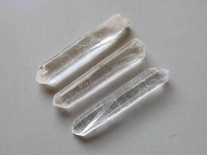 Peut inclure: Trois pointes de cristal de quartz transparentes, chacune avec un sommet naturel et brut et une extr&eacute;mit&eacute; polie et pointue. Les cristaux sont transparents avec de subtiles inclusions internes, dispos&eacute;s en diagonale.