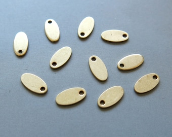 May include: A collection of small, oval-shaped brass tags with a single hole at one end. The tags are scattered on a gray surface. These tags are likely used for jewelry making or crafting.