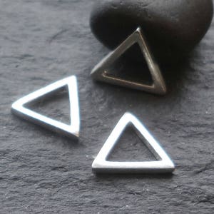 May include: Three silver-coloured, open-frame triangle charms are displayed on a dark, textured surface. The geometric shapes are evenly spaced, with one slightly out of focus. A dark, rounded stone is in the background.