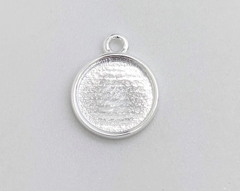 Può includere: Un ciondolo rotondo color argento con un anello nella parte superiore. Il ciondolo ha un'area incassata per aggiungere un disegno o un'immagine. Il ciondolo è mostrato su uno sfondo grigio chiaro.