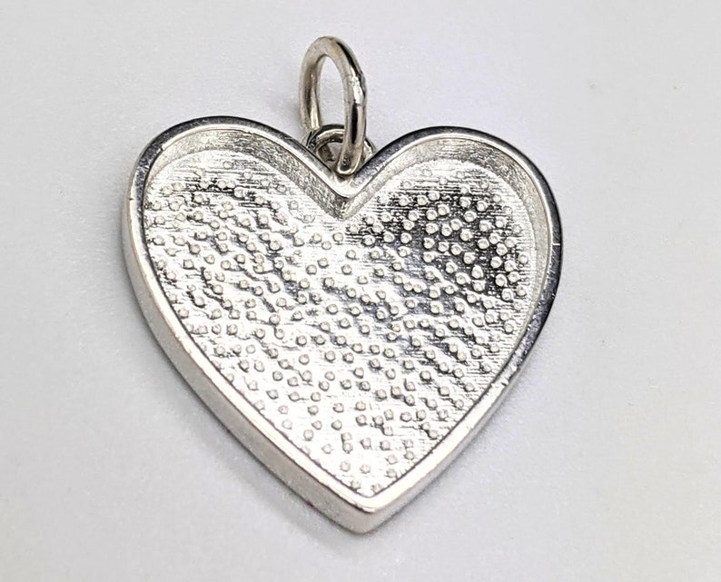 May include: Silver heart-shaped pendant with a textured surface. The pendant has a loop at the top for attaching a chain.