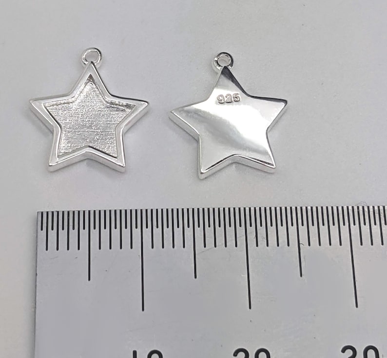 Może przedstawiać: Dwa srebrne charms w kształcie gwiazdy z k&oacute;łkiem do zawieszania na biżuterii. Charms mają około 2,5 cm średnicy i są oznaczone znakiem 925.