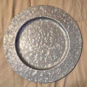 Puede incluir: Un plato de carga redondo de metal galvanizado con una superficie texturizada. El plato tiene un borde ancho y un centro ligeramente elevado. El metal tiene un color plateado con un aspecto moteado.