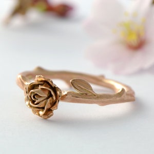May include: A rose gold ring with a delicate rose and leaf design. The ring is made to look like a branch with a rose growing from it.