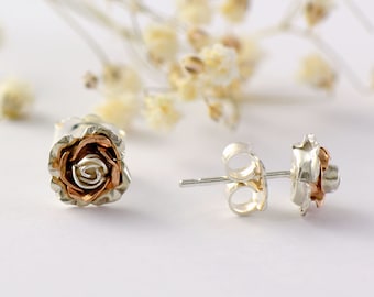 Puede incluir: Un par de pendientes de plata con forma de rosa y un centro de oro rosa. Los pendientes se muestran sobre un fondo blanco con flores secas.