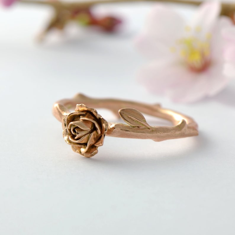 May include: A rose gold ring with a delicate rose and leaf design. The ring is made to look like a branch with a rose growing from it.