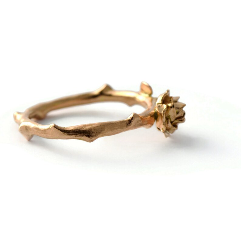 May include: A gold ring with a delicate, twig-like band and a small flower design. The ring is made of metal and has a smooth, polished finish.