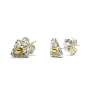 May include: A pair of silver stud earrings in a triangular shape. The earrings have a textured, uneven edge and a small, gold-coloured ball in the centre. The earrings are shown against a white backdrop.