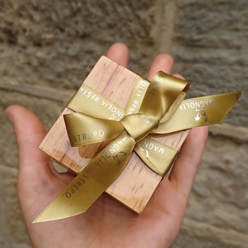 May include: A small wooden box tied with a gold ribbon. The ribbon has the text "Magnolia Strepo" printed on it.