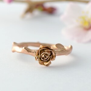 May include: A rose gold ring with a rose design on a twig-like band.