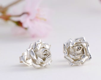 Puede incluir: Un par de pendientes de botón de plata con forma de rosa. Las rosas son detalladas y realistas, con pétalos que parecen estar hechos de metal.