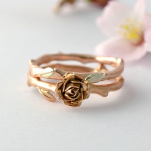 Puede incluir: Un anillo de oro rosa con un dise&ntilde;o de rosa y dos bandas. Las bandas est&aacute;n hechas para parecer ramas con hojas.