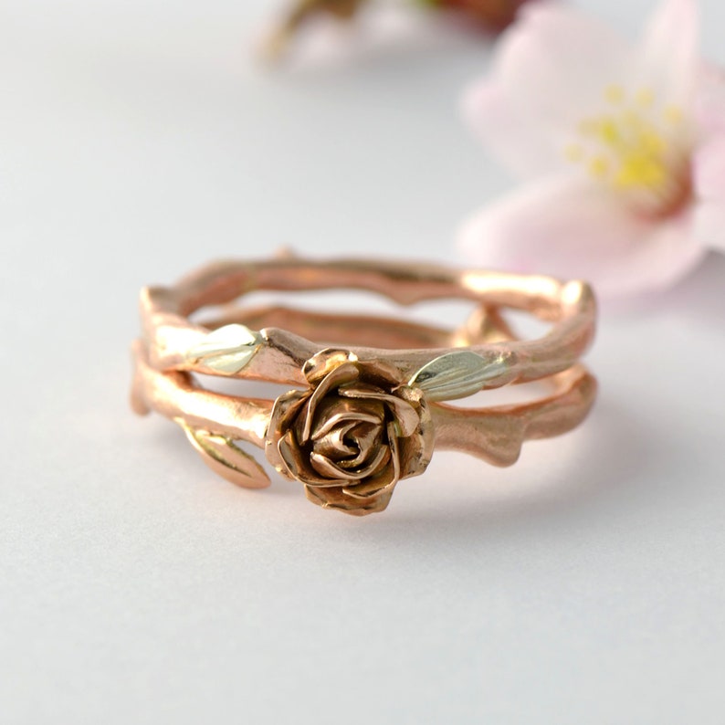 Puede incluir: Un anillo de oro rosa con un dise&ntilde;o de rosa y dos bandas. Las bandas est&aacute;n hechas para parecer ramas con hojas.