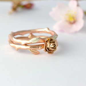 May include: A rose gold ring with a rose and leaves design. The ring is made of two bands, one with a thicker stem and the other with a thinner stem. The rose is in the center of the ring and is surrounded by leaves.