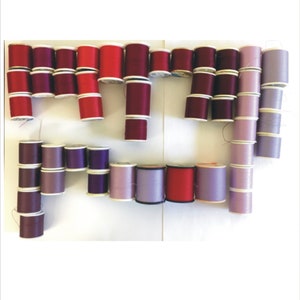 May include: A collection of spools of thread in various shades of red, purple, and lavender. The spools are arranged in a staggered pattern on a white surface.
