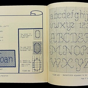May include: Open book pages with cross-stitch instructions. One page features a scroll design with text and a stitched "by Joan" sample. The opposite page displays a grid with the alphabet and numbers for backstitch.