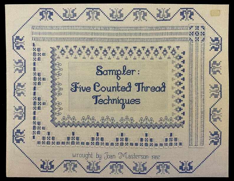 May include: A hand-stitched sampler with blue and white thread on a cream-colored background. The text reads "Sampler: Five Counted Thread Techniques" and "wrought by Joan Masterson 1982". The sampler features various counted thread patterns.