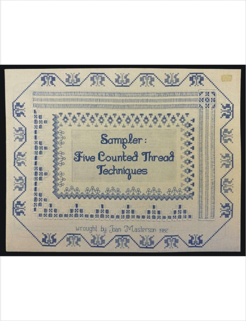 May include: A framed textile sampler with blue embroidery on a cream background. The sampler features the text "Sampler: Five Counted Thread Techniques" and is framed with a decorative border. The text "wrought by Joan Masterson 1982" is also included.