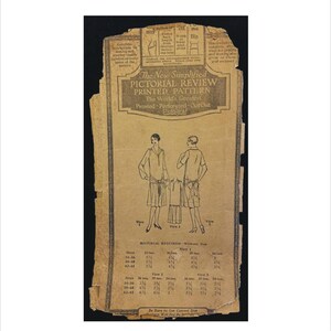 May include: An aged, tan-colored paper pattern with the text "The New Simplified Pictorial Review Printed Pattern." The pattern features illustrations of two women's dresses and size charts. The paper is torn and worn.