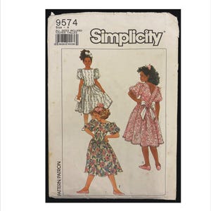 May include: A vintage Simplicity sewing pattern with illustrations of three dresses. The pattern includes the number 9574 and the word "Simplicity". The dresses are in white, pink, and floral patterns. The pattern is for size A.