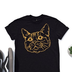 Puede incluir: Camiseta negra con un contorno dorado de la cara de un gato.