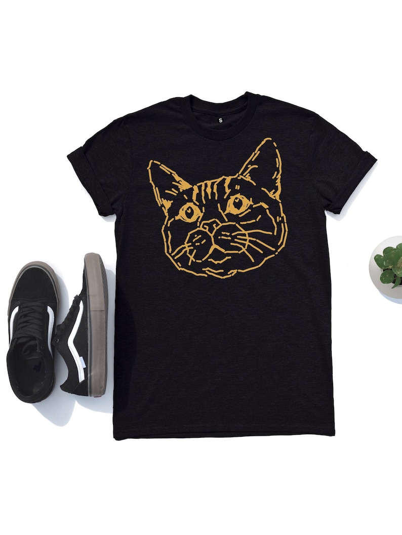 Puede incluir: Camiseta negra con un contorno dorado de la cara de un gato.