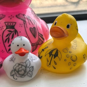 Puede incluir: Una colección de patitos de goma de varios colores. El pato blanco tiene un tatuaje de rosa, el pato amarillo tiene un tatuaje de paraguas y el pato rosa tiene un tatuaje de mano. El pato amarillo tiene la palabra "CLEAN" impresa.