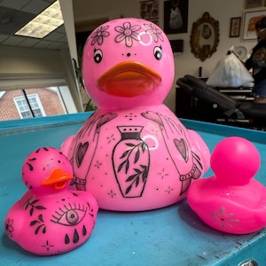 Puede incluir: Una colección de patos de goma rosas con diseños de tatuajes negros. El pato más grande presenta acentos florales en la cabeza, un pico rojo y un diseño de jarrón en el pecho. Los patos más pequeños tienen diseños de ojos y flores. Los patos están sobre una superficie azul.