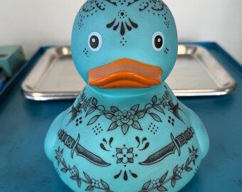 Puede incluir: Un pato de goma turquesa con diseños de tatuajes florales y de daga negros. El pato tiene un pico naranja y ojos blancos. El pato está sobre una superficie azul.