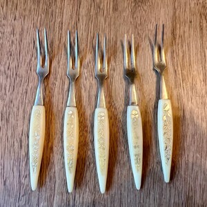 May include: Five vintage dessert forks with cream-colored handles and silver tines. The handles are decorated with floral and leaf patterns. The forks are arranged on a wooden surface.