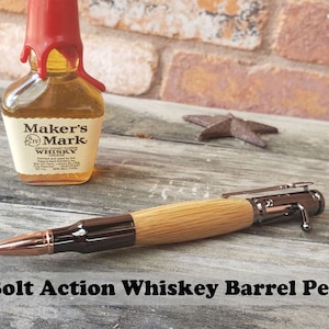 May include: A wooden pen with a metal bolt action mechanism and a copper bullet tip. The pen is resting on a wooden surface with a miniature bottle of Maker's Mark whiskey in the background.