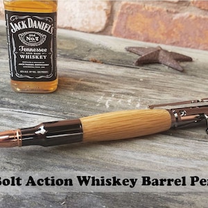 May include: A wooden pen with a metal bolt action mechanism and a copper bullet tip. The pen is made from a whiskey barrel and has a black finish. The pen is sitting on a wooden surface with a bottle of Jack Daniel's Tennessee Whiskey in the background.