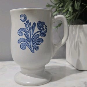 Peut inclure: Mug en céramique blanche avec un motif floral bleu. La tasse a une base en piédestal et une anse incurvée. Le motif floral bleu est un dessin stylisé de fleurs et de feuilles. La tasse est probablement pour le café ou le thé.