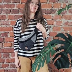 May include: A woman wearing a black and white striped long-sleeved shirt and brown pants is standing in front of a brick wall. She is wearing a gray and black fanny pack with the text "hai tao" on it. A large green leaf plant is to the right of the woman.
