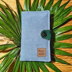 Op de afbeelding: Een lichtblauwe stoffen portemonnee met een groene sluiting en een bruine knoopsluiting. De portemonnee heeft een bruine leren patch met de tekst "hai roo". De portemonnee ligt op een houten oppervlak met groene bladeren.