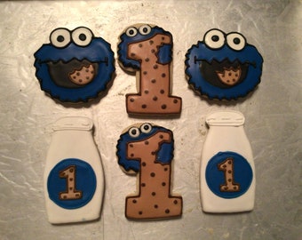 May include: Six decorated sugar cookies, two blue cookies with a cookie monster face and the number one, two brown cookies with the number one and brown dots, and two white cookies with a blue circle and the number one.